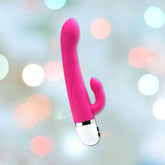The VeDO Wink Rabbit Vibe in Hot Pink, by VeDO, is a dual-ended vibrator featuring a G-Spot and Clitoris targeted design against a backdrop of blurred multicolored lights. It has a white handle with a silver accent and offers twelve gratifying modes to explore.